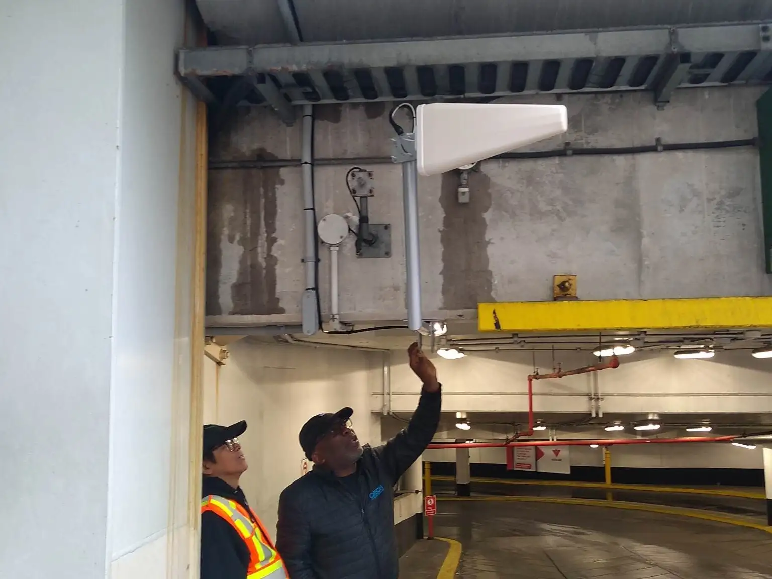 Two technicians in a parkade checking a mounted indoor antenna above the entry ramp.